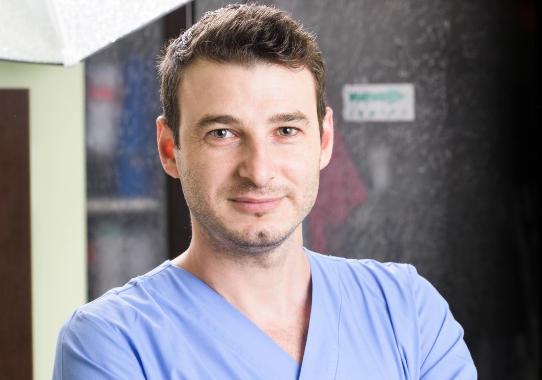 Bărbat cu părul brun, îmbrăcat în halat medical albastru, zâmbind în fața camerei.