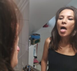 Femeie cu părul lung, îmbrăcată în negru, care își scoate limba în fața oglinzii.