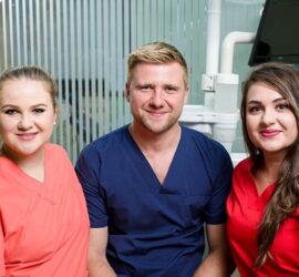 Trei medici dentiști zâmbind în uniformă, în interiorul unei clinici dentare.