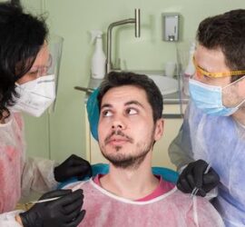 Un pacient în scaunul stomatologic discutând cu doi medici dentiști în echipament de protecție.