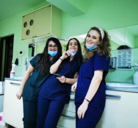 Trei asistente medicale zâmbind în uniformă, cu măști, într-un mediu de lucru.