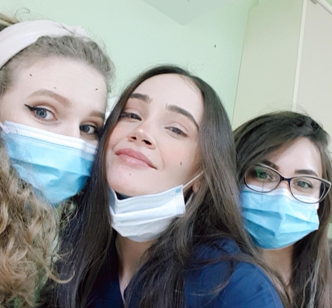 Trei femei zâmbind, fiecare purtând o mască de protecție albastră, în interior.