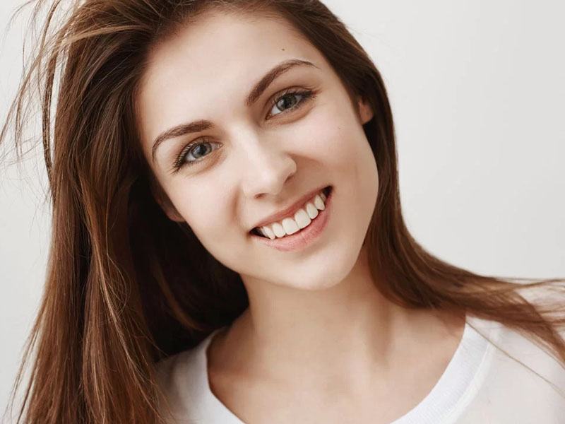 Tânără cu părul lung și zâmbet, purtând un tricou alb, pe un fundal neutru.