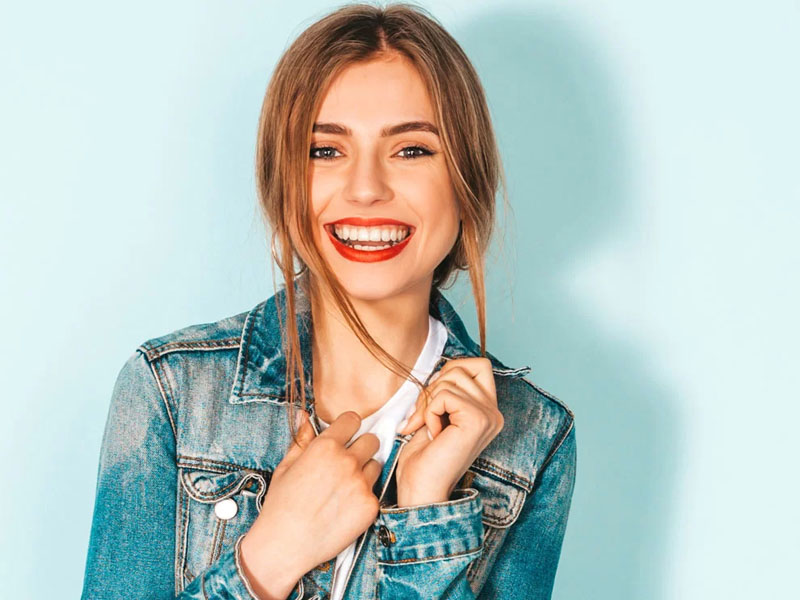 Tânără cu păr lung și blond, zâmbind, îmbrăcată într-o jachetă din denim.