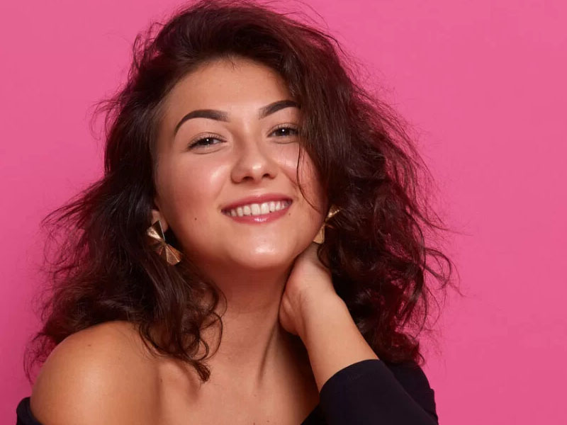 Fată cu păr ondulat și zâmbet, purtând cercei, pe un fundal roz.