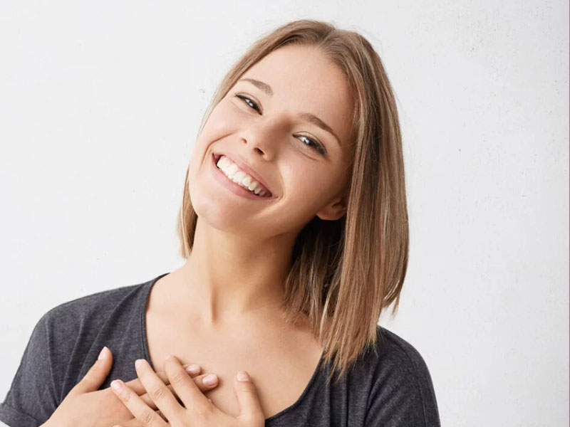 Tânără cu părul scurt, zâmbind și cu mâinile pe piept, pe un fundal deschis.