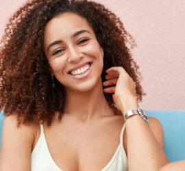 Fată cu păr creț și zâmbet, purtând un tricou deschis la culoare și brățară pe încheietura mâinii.