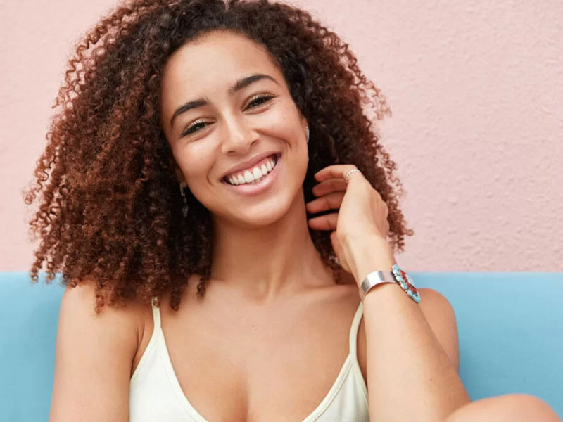 Fată cu păr creț și zâmbet, purtând un tricou deschis la culoare și brățară pe încheietura mâinii.