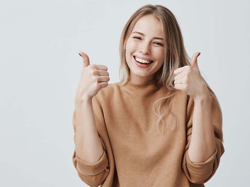 Femeie tânără cu păr lung, zâmbind și arătând degetele mari ridicate în semn de aprobat.