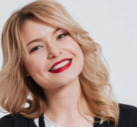 Femeie cu păr blond ondulat și buze vopsite în roșu, zâmbind la cameră.