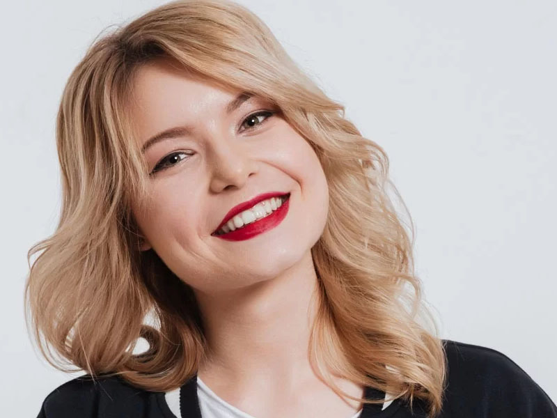 Femeie cu păr blond ondulat și buze vopsite în roșu, zâmbind la cameră.