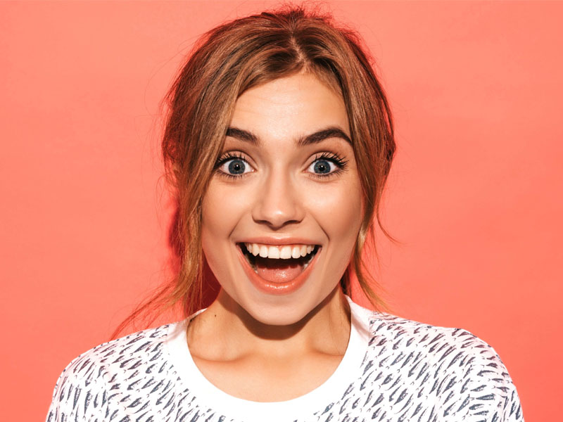 Femeie cu părul lung și blond, zâmbind larg pe un fundal roz.