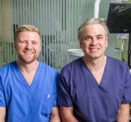 Doi medici îmbrăcați în uniforme albastre și mov, zâmbind în fața camerei într-un mediu medical.