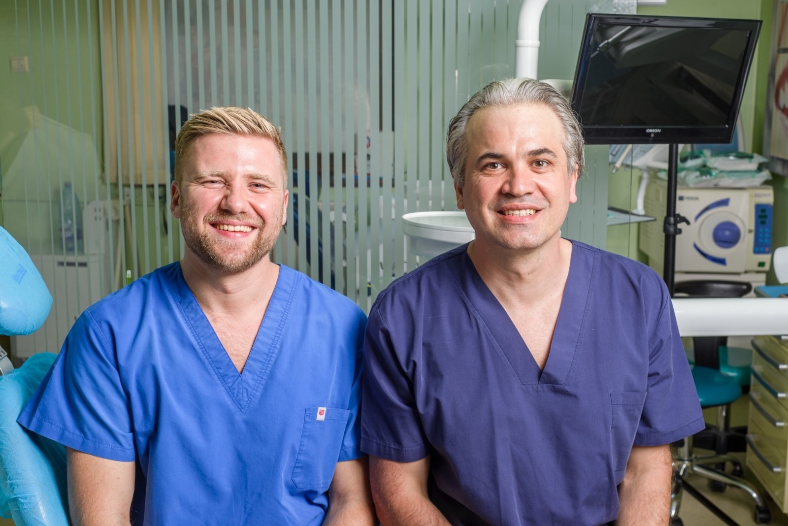 Doi medici îmbrăcați în uniforme albastre și mov, zâmbind în fața camerei într-un mediu medical.