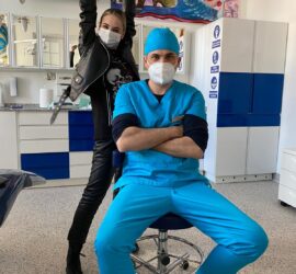Un medic stomatolog îmbrăcat în uniformă albastră stând pe un scaun, cu o pacientă în spate care ridică brațele.