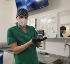 Medic îmbrăcat în uniformă verde, cu mască și mănuși, ținând un recipient în mână.