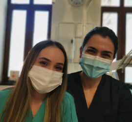 Două asistente medicale zâmbind, purtând măști și uniforme de lucru, într-un mediu medical.