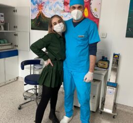 Două persoane, una îmbrăcată în uniformă medicală albastră și cealaltă într-o rochie verde, ambele purtând măști.