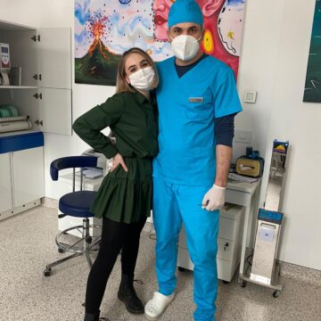 Două persoane, una îmbrăcată în uniformă medicală albastră și cealaltă într-o rochie verde, ambele purtând măști.