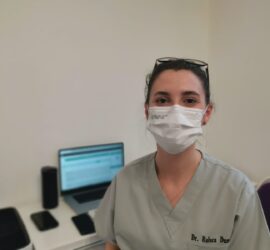 Doctor purtând mască și uniformă medicală, stând lângă un birou cu un computer.