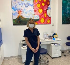 Asistent medical purtând o uniformă albastră și mască, stând în fața unui tablou colorat.
