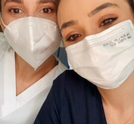Două asistente medicale zâmbind, purtând măști de protecție și uniforme de lucru.