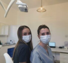Două asistente dentare purtând măști, zâmbind în fața camerei, în interiorul unui cabinet stomatologic.