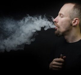 Bărbat cu barbă care expiră vapori dintr-un dispozitiv de vaping într-un fundal întunecat.