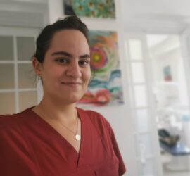 Femeie cu păr lung, îmbrăcată în uniformă medicală roșie, zâmbind în interiorul unei clinici.