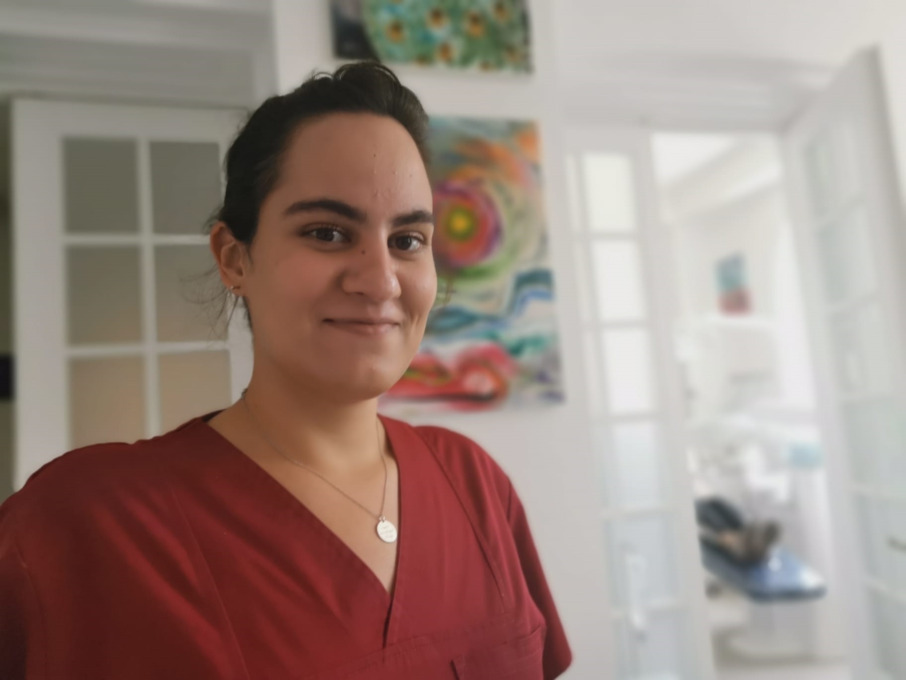 Femeie cu păr lung, îmbrăcată în uniformă medicală roșie, zâmbind în interiorul unei clinici.