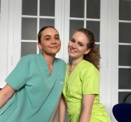 Două asistente medicale zâmbind, purtând uniforme verzi și galbene, într-un mediu interior.