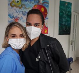 Două femei zâmbind și purtând măști de protecție, una îmbrăcată în uniformă medicală.