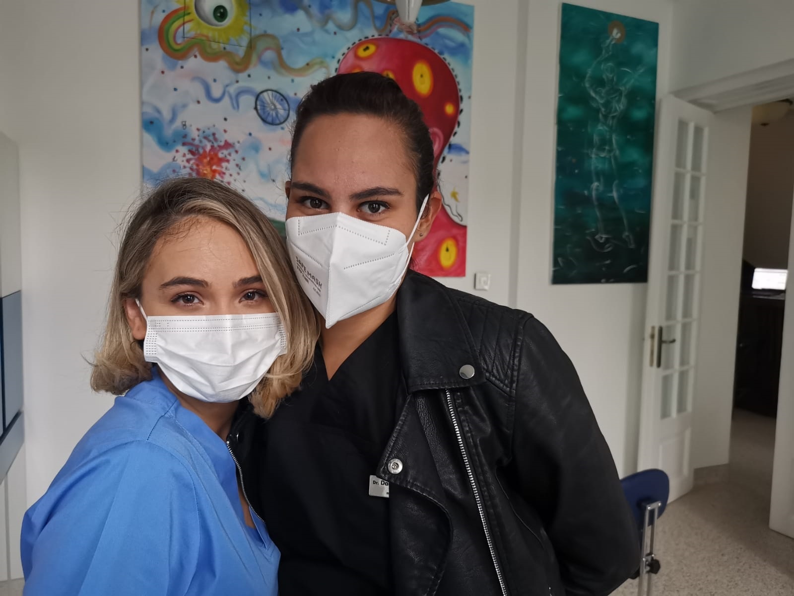 Două femei zâmbind și purtând măști de protecție, una îmbrăcată în uniformă medicală.