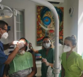Un medic stomatolog examinează un pacient în cabinet, cu doi asistenți în echipament de protecție.