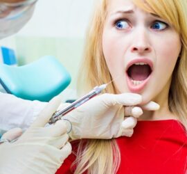 Femeie cu părul blond și lung, având gura deschisă, primind o injecție de anestezie locală la dentist.