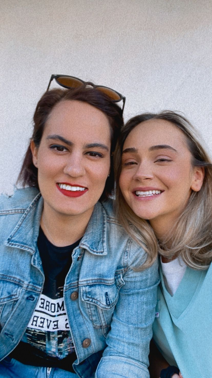 Două femei zâmbitoare, una cu o bluză verde și cealaltă cu o jachetă de blugi, pozând împreună.