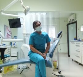 Asistentă medicală purtând mască, îmbrăcată în uniformă albastră, stând pe un scaun dintr-un cabinet stomatologic.