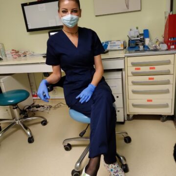 Asistent medical purtând uniformă albastră, mănuși și mască, așezat pe un scaun.