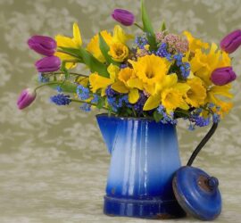 Buchet de flori colorate, inclusiv lalele și narcise, într-o cană albastră de metal.