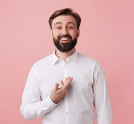 Bărbat tânăr cu barbă și zâmbet larg, îmbrăcat în cămașă albă, pozând pe un fundal roz.