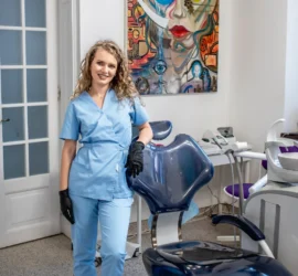 Femeie blonda zambeste inmbracata in uniforma medicala sprijinita de fotoliul dentar