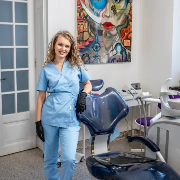 Femeie blonda zambeste inmbracata in uniforma medicala sprijinita de fotoliul dentar