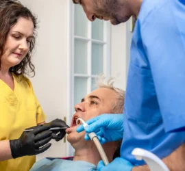 Femeie medic stomatolog in uniforma galbena se uita ]n cavitatea bucala a unui pacient carunt aflat pe fotoliul stomatologic